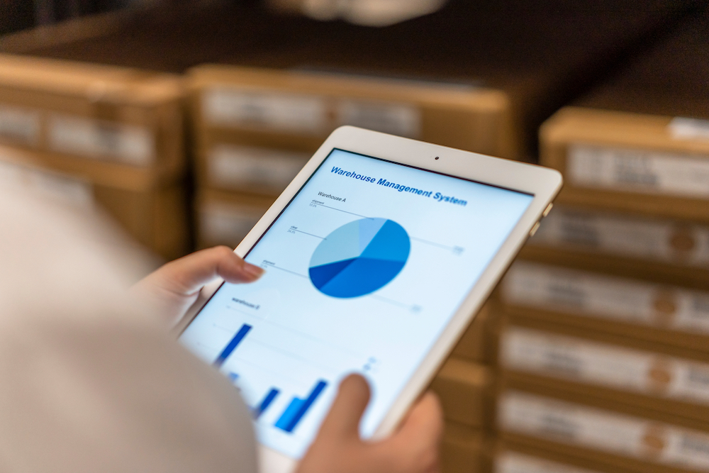 A person stands in a warehouse holding a tablet that is displaying inventory status through charts and graphs. Smart inventory uses the power of data and analytics combined with machine learning to help manufacturing organizations accurately plan for when and where their products will be needed.