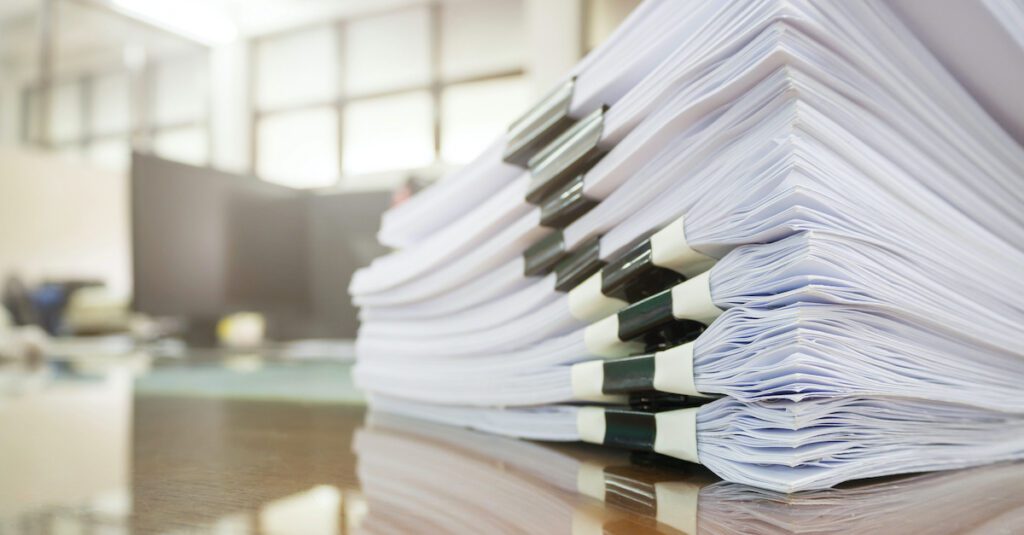A stack of documents represents how the volume of documentation that flows through a financial services or banking organization can cause a slowdown in the process and negatively impact customer experience.