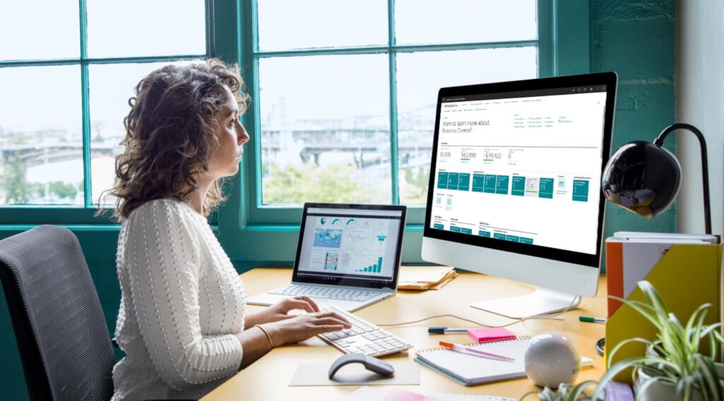 Business-Central-Wave-2-2022 Feature Image, a woman sits at her desk using Microsoft 365 business central on her computer